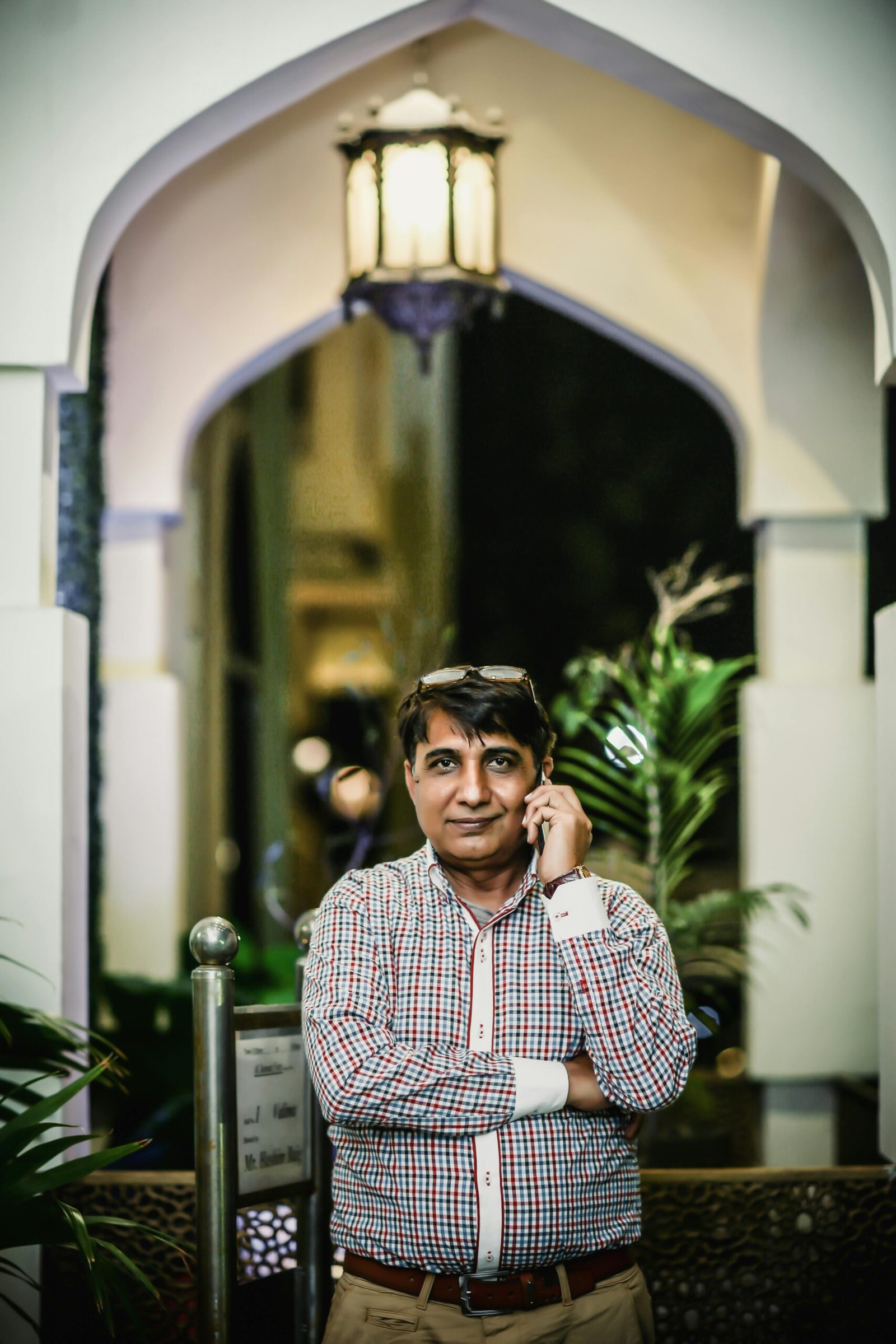 A South Asian man in casual attire talks on his mobile phone under a lit archway.